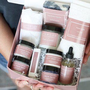 Pu&ograve; includere: Una scatola regalo rosa piena di prodotti per il bagno e il corpo, tra cui una candela, sapone, lozione per il corpo, scrub allo zucchero, maschera per il viso, sali da bagno e olio per il corpo. Tutti i prodotti sono etichettati con il nome "Lynwood Candle Company".