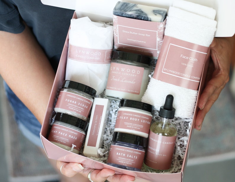 Pu&ograve; includere: Una scatola regalo rosa piena di prodotti per il bagno e il corpo, tra cui una candela, sapone, lozione per il corpo, scrub allo zucchero, maschera per il viso, sali da bagno e olio per il corpo. Tutti i prodotti sono etichettati con il nome "Lynwood Candle Company".
