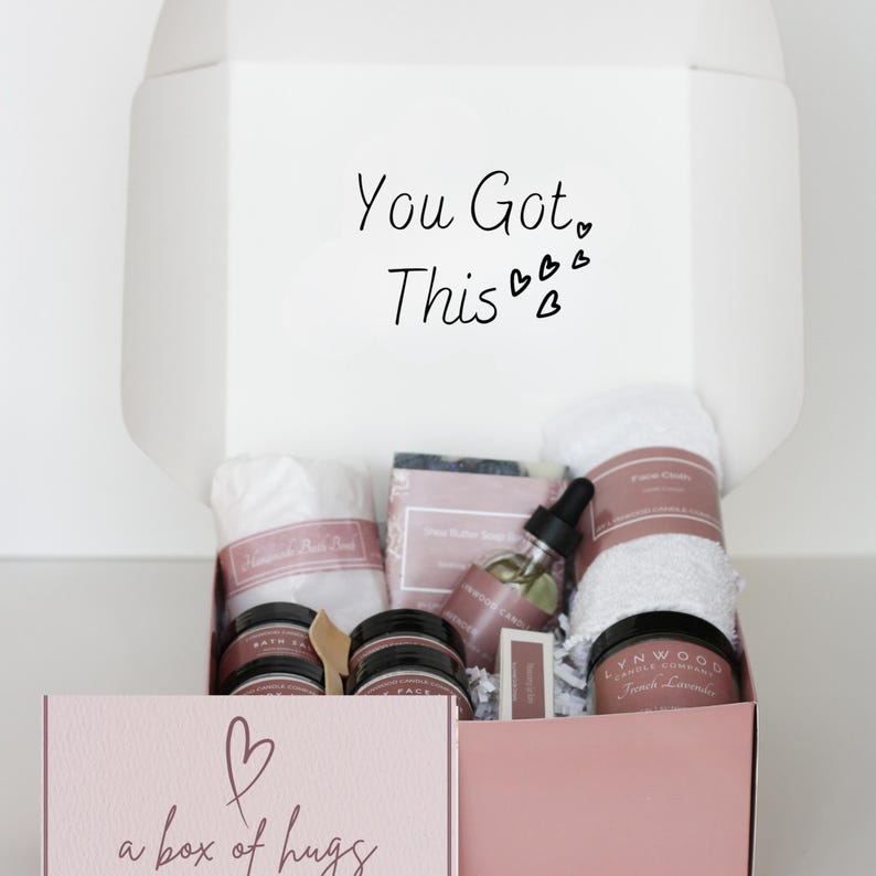 Pu&ograve; includere: Una scatola regalo rosa con il testo "You Got This" e un design a forma di cuore. La scatola &egrave; piena di prodotti per il bagno e il corpo, tra cui un asciugamano bianco, una bottiglia di olio e una candela. La scatola ha anche il testo "a box of hugs" sul davanti.