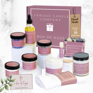 May include: A gift box labeled "Box of Hugs" filled with self-care items. Includes jars of bath salts, face mask, sugar scrub, body lotion, and a French lavender candle. Also features a shea butter soap bar, lip balm, and a small white towel.