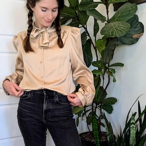 Op de afbeelding: Een vrouw draagt een lichtbruin satijnen blouse met een strik, een zwarte jeans en bruine enkellaarsjes. Ze staat voor een kamerplant en een gitaar.