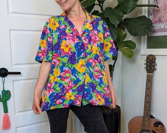 Könnte beinhalten: Eine Frau trägt ein kurzärmliges Hemd mit blauem, rosa, gelbem und grünem Blumendruck und eine schwarze, kurze Hose.