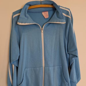 Könnte beinhalten: Hellblaue Trainingsjacke mit Reißverschluss und weißen Streifen an Ärmeln und Kragen. Die Jacke hat einen durchgehenden Reißverschluss, zwei Vordertaschen und ein Etikett am inneren Kragen. Die Jacke hängt an einem Holzkleiderbügel.