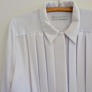 Puede incluir: Una blusa blanca de manga larga con cuello clásico y parte delantera plisada. La etiqueta dice "VALERIE STEVENS". La blusa está colgada en una percha de madera.