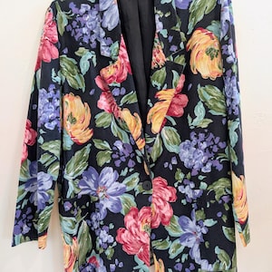 Puede incluir: Un blazer negro con un vibrante estampado floral con flores rosas, amarillas y moradas. El blazer tiene un cierre de un solo botón y dos bolsillos delanteros. La chaqueta está colgada en una percha de madera.