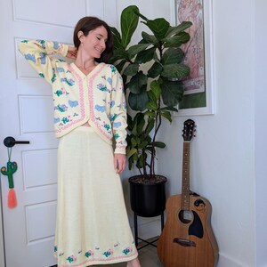 Könnte beinhalten: Eine Frau trägt ein gelbes Strick-Cardigan und einen Rock im Set mit Blumenstickerei. Das Cardigan hat einen V-Ausschnitt und Knöpfe vorne. Der Rock ist lang und fließend. Die Frau steht vor einer Topfpflanze und einer Gitarre.