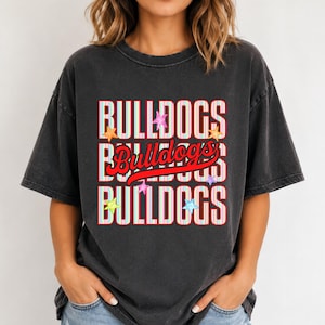 K&ouml;nnte beinhalten: Ein anthrazitfarbenes Kurzarm-T-Shirt mit einem Grafikdruck, der das Wort BULLDOGS in einer gestreiften Schrift wiederholt, &uuml;berlagert mit einem roten Schreibschrift-Wort Bulldogs und mehreren kleinen bunten Sternen im Design.