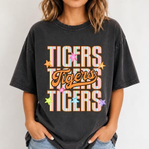 K&ouml;nnte beinhalten: Ein anthrazitfarbenes Kurzarm-T-Shirt mit dem dreimal vertikal untereinander gedruckten Wort TIGERS. Die obere und untere Zeile sind in Blockschrift mit vertikalen Streifen gehalten, w&auml;hrend die mittlere Zeile in orangefarbener Schreibschrift steht.