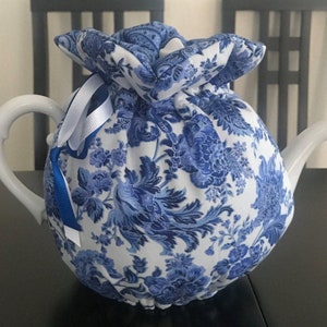 May include: A white ceramic teapot with a blue and white floral patterned tea cozy. The cozy is tied with white and blue ribbons. The teapot has a curved handle and spout. The tea cozy is made of fabric.