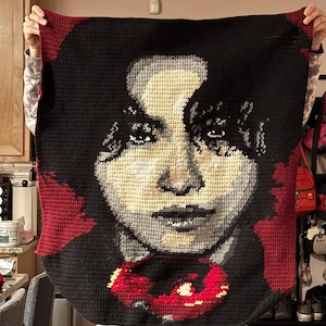 May include: A crocheted blanket featuring a portrait of a person with black hair and a red flower. The blanket is made with black, white, red, and gray yarn.
