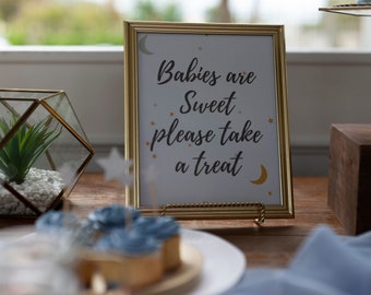 Puede incluir: Un cartel con marco dorado, fondo blanco y texto negro que dice "Babies are Sweet please take a treat". El cartel está decorado con estrellas y lunas doradas.