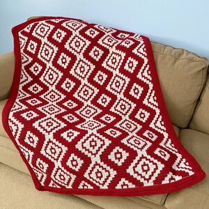 May include: A red and white crocheted throw blanket with a geometric diamond pattern. The blanket is folded and draped over a brown couch.