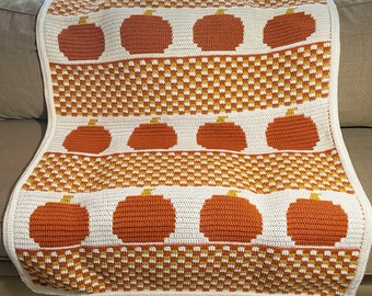 May include: A crocheted throw blanket with a white background and orange pumpkins. The blanket has a repeating pattern of pumpkins and a checkered design.