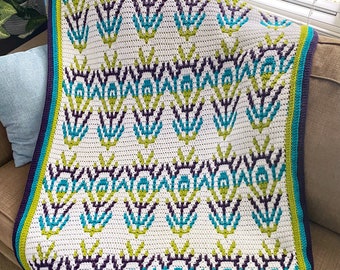 May include: A white crocheted throw blanket with a geometric pattern in shades of teal, green, and purple. The blanket has a crocheted border in teal, green, and purple.