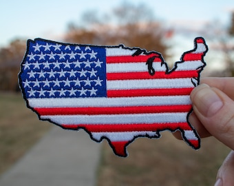 May include: A red, white, and blue embroidered patch shaped like the United States of America. The patch is made of fabric and has a stitched outline.