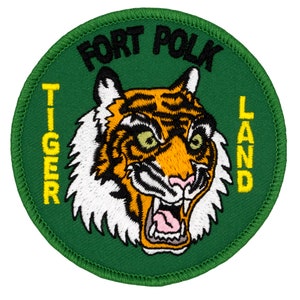 May include: A green circular patch with yellow text that reads "Tiger Land" and "Fort Polk" in black text. A cartoon tiger with orange and black stripes and a white mane is in the center of the patch.