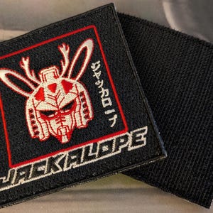 May include: Black square patch with a red and white robotic jackalope design. The patch features the word "JACKALOPE" in white block letters and Japanese text. The patch has a hook and loop backing.
