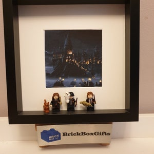 May include: A framed LEGO minifigure display featuring Harry Potter, Ron Weasley, and Hermione Granger with Hedwig the owl. The background image shows Hogwarts castle at night. The frame is black with a white mount and the bottom of the frame has a small logo that says "BrickBoxGifts".