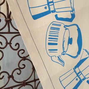 May include: A white fabric kitchen towel with a blue design of different coffee makers. The design includes a traditional stovetop espresso maker, a percolator, and a French press.