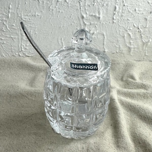 May include: A clear, cut-glass sugar bowl with a lid and a small spoon. The bowl has a diamond pattern and the word "Shannon" is on a label on the lid. The spoon is inserted into the bowl.