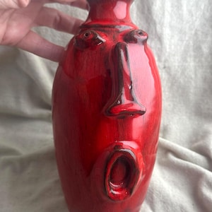 May include: A glossy red ceramic vase featuring a sculpted face with a prominent nose and an open, oval-shaped mouth. The surface has a deep, vibrant red glaze with darker red shading and a smooth, reflective finish on the rounded body.
