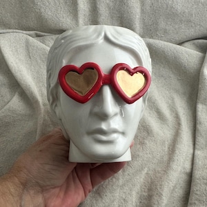 May include: A white ceramic head sculpture wearing red heart-shaped sunglasses with gold lenses. The sculpture has classical features and is held by a hand.