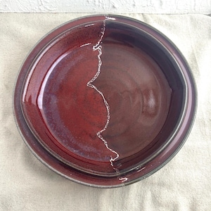 May include: Two ceramic plates in red and brown tones. The plates are round with a raised rim and a glossy finish. A white, jagged line divides the plates, creating a unique design. These plates are likely for serving food.