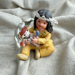 May include: A ceramic figurine of a Native American child in a yellow outfit, holding a doll. The child has black hair, a white headband with red dots, and a necklace. The doll has a red and white headdress. The figurine is on a beige surface.