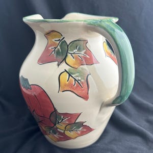 May include: Cream-colored ceramic pitcher with a green handle and rim. The pitcher is decorated with colorful autumn leaves in red, yellow, and green. The pitcher has a wide base and a narrow spout.