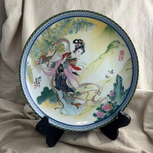 May include: A decorative porcelain plate featuring a central illustration of a figure in traditional attire holding a folding fan. The scene includes banana leaves, pink flowers, and a mountain landscape. The plate has a blue and gold patterned border.