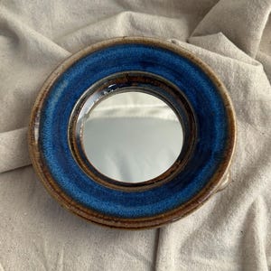 May include: A circular ceramic mirror featuring a vibrant blue glazed rim and a brown edge. The mirror's reflective surface is framed by the blue and brown ceramic. This is a decorative item.