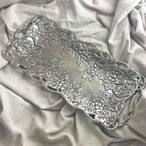 May include: A rectangular silver-colored serving tray with a grape and leaf design around the edges. The tray has a smooth, reflective surface in the center and handles on each end. The tray is made of metal and is suitable for serving food or displaying items.