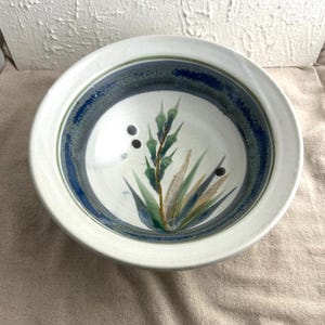 May include: A round, white ceramic bowl with a wide rim. The interior features a blue and green painted design with a central botanical illustration in shades of green, blue, and tan. Three small holes are visible.