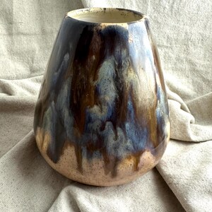 May include: A handcrafted ceramic vase with a wide base and a narrow opening. The vase features a blend of colors, including brown, blue, and beige, with a glossy finish. The glaze drips down the sides, creating a unique, artistic look. This decorative vase is suitable for home decor.