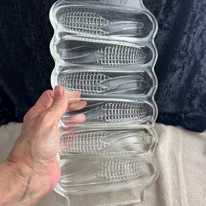 May include: A clear glass dish with five indentations shaped like corn on the cob. The dish is designed to hold corn on the cob.