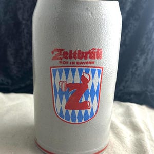 May include: A white ceramic beer stein with a red and blue logo that says "Zeltbrauerei Hof in Bayern". The logo features a person holding a beer stein and a large letter "Z" in the center.