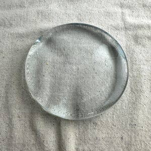 May include: A clear, circular glass object with a slightly textured surface. The transparent disc is round and appears to be a coaster or small decorative piece. The glass has a subtle, rippled edge.