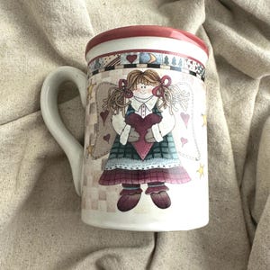 May include: White ceramic mug featuring a whimsical angel design. The angel holds a heart and wears a plaid dress with wings. The rim is red, and the handle is white. The mug is decorated with hearts and stars.