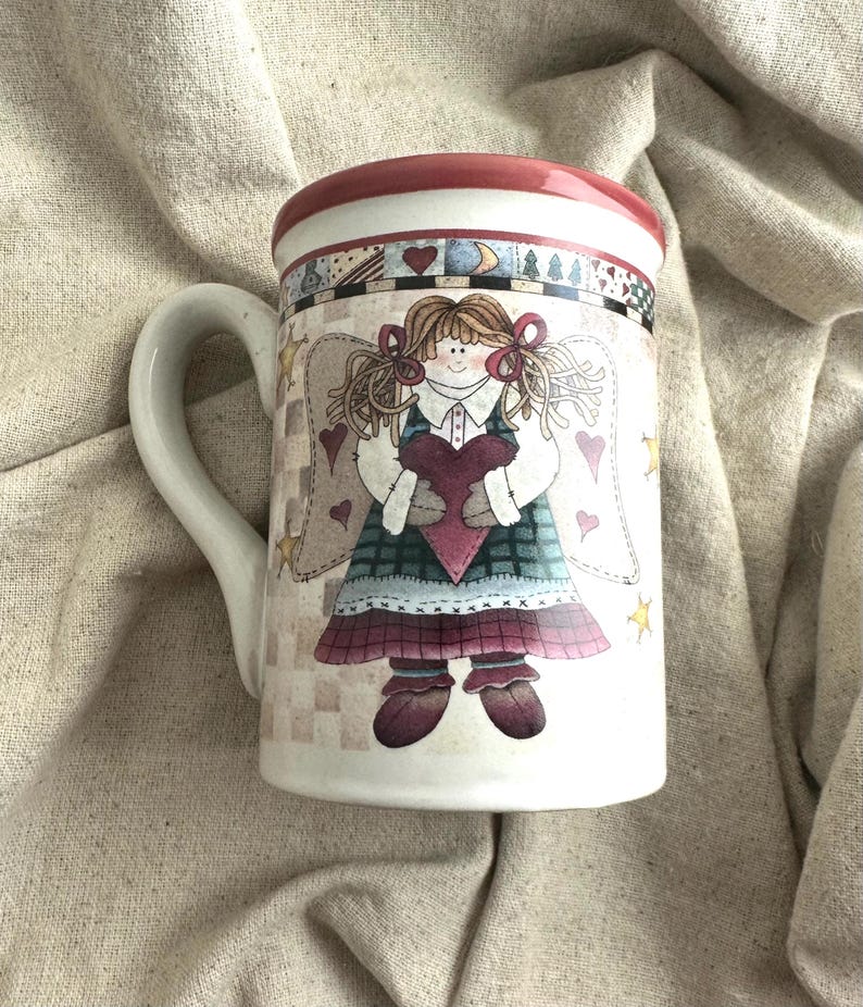 May include: White ceramic mug featuring a whimsical angel design. The angel holds a heart and wears a plaid dress with wings. The rim is red, and the handle is white. The mug is decorated with hearts and stars.