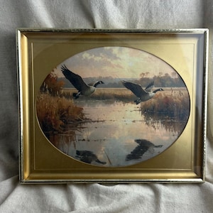 May include: A rectangular framed print featuring an oval-shaped landscape painting of two Canada geese in flight over a marshy river at sunset. The artwork is encased in a metallic gold frame with a matching gold matting surrounding the oval image.