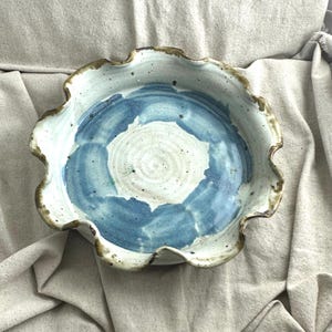May include: A handmade ceramic pie dish with a ruffled edge. The interior features a blue and white abstract design. The rim has a brown trim. The dish is set against a neutral-coloured fabric background.