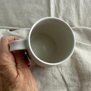 Op de afbeelding: Een witte keramische mok met een gebogen oor. De mok wordt door een hand vastgehouden, waarbij de binnenkant en het ontwerp van het oor te zien zijn. De binnenkant van de mok is lichtbeige.