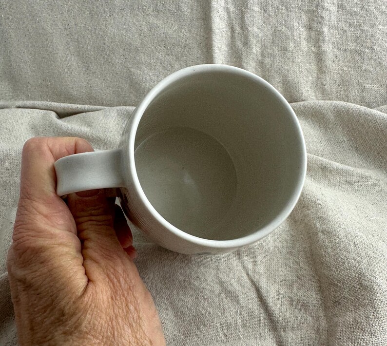 Op de afbeelding: Een witte keramische mok met een gebogen oor. De mok wordt door een hand vastgehouden, waarbij de binnenkant en het ontwerp van het oor te zien zijn. De binnenkant van de mok is lichtbeige.