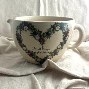 May include: Cream-colored ceramic mixing bowl with a handle and spout. A heart-shaped wreath of plums, berries, and white flowers decorates the front. The text "In all things give thanks" is printed below the wreath.