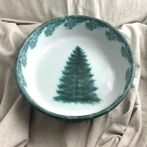 May include: A white ceramic bowl with a teal green rim and a teal green fir tree design in the center. The bowl has a simple, rustic design, perfect for serving food or as a decorative piece.