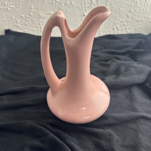 May include: A pink ceramic vase with a unique handle and a curved spout. The vase has a rounded base and a smooth, glossy finish. The handle curves gracefully from the base to the top, creating an elegant silhouette.