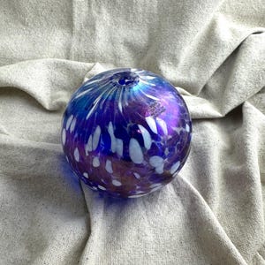May include: A spherical glass art piece with a deep blue and purple color scheme, speckled with white accents. The glass has a glossy finish and a small opening at the top. The text "KEEP OUT OF REACH OF CHILDREN" is visible on a small label.