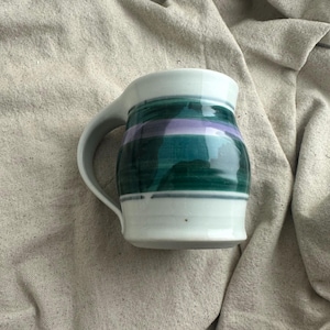 May include: A handmade ceramic mug with a white base and rim. The mug features a green and purple stripe design around the middle. The handle is white and curved.
