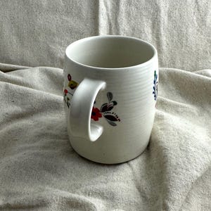Op de afbeelding: Witte keramische mok met een gebogen handvat, versierd met bloemmotieven in rood, blauw en geel. De mok heeft een getextureerd oppervlak en een brede opening, geschikt voor warme dranken. Het bloemendesign voegt kleur toe.
