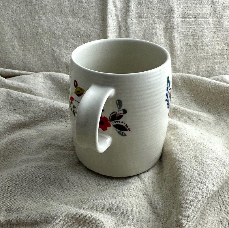 Op de afbeelding: Witte keramische mok met een gebogen handvat, versierd met bloemmotieven in rood, blauw en geel. De mok heeft een getextureerd oppervlak en een brede opening, geschikt voor warme dranken. Het bloemendesign voegt kleur toe.
